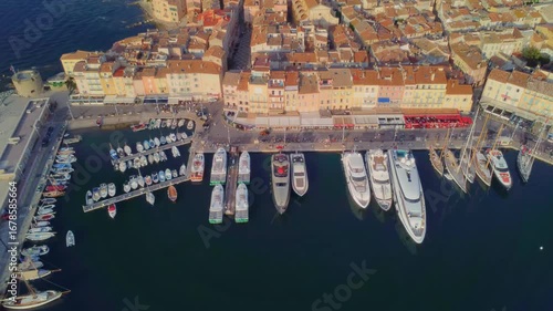 aerial drone shot capturing the iconic Port of Saint Tropez on the French Riviera, showcasing a vibrant array of luxury yachts and traditional fishing boats moored alongside the charming, historic tow