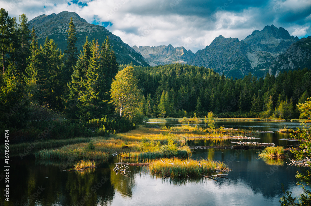 Fototapeta premium As the sun sinks behind the towering peaks, the quiet lake reflects the sky in shades of orange and gold, offering a moment of untouched silence in the heart of the mountains.