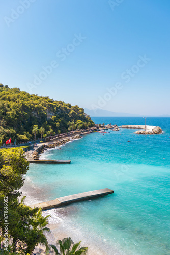 The Aegean coast on a sunny summer day in Kusadasi. Azure water, piers, green hills, palm trees and a promenade with the flag of Turkey. A picturesque seascape, a popular tourist destination for