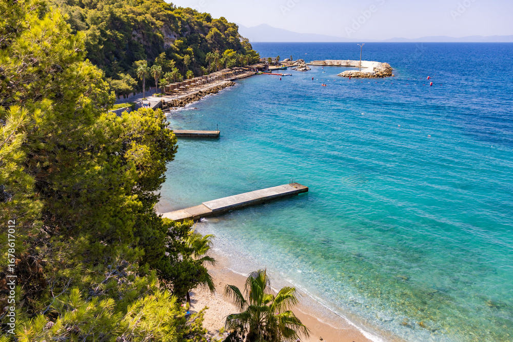 Obraz premium The Aegean coast on a sunny summer day in Kusadasi. Azure water, piers, green hills, palm trees and a promenade with the flag of Turkey. A picturesque seascape, a popular tourist destination for