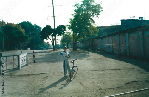 Ten-year-old boy with bicycle stands near warehouses and railway crossing in Odessa, captured in film photography style from mid-1990s in Ukraine