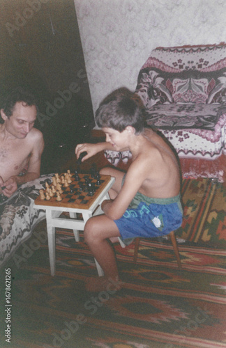 Father and ten-year-old son playing chess in home room during summer vacation, captured in film photography style from early 1990s in Ukraine