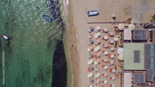 aerial drone shot captures the vibrant and luxurious beach scene of Saint Tropez, France, showcasing rows of beach umbrellas, sun loungers, and crystal clear turquoise waters with boats docked nearby.