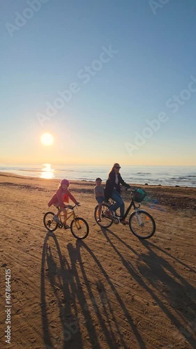 family with children riding with bicycles on beach sand at sunset