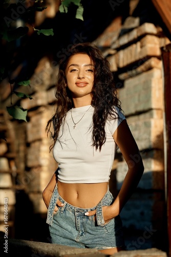 Smiling young woman in a white top and denim shorts poses outdoors