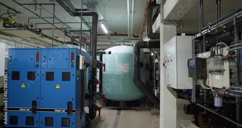 Inside the plant room of a public aquatic center. This shot shows the complex water filtration and circulation system, including large sand filters, pumps, pipes, and electronic control panels.