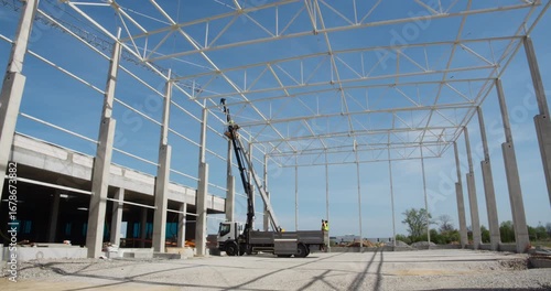 Team of construction workers collaborates on erecting the steel frame of a warehouse. One worker in a cherry picker (boom lift) guides a roof truss into place, assisted by the ground crew.
