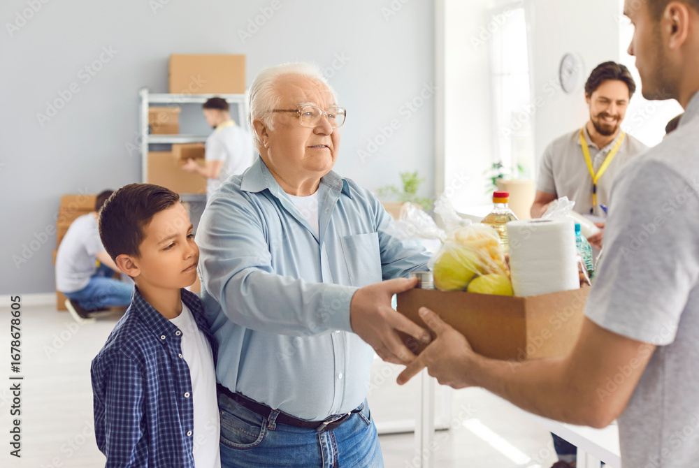 Fototapeta premium Sad elderly man and child receive a box of food from a volunteer, close-up. Group of volunteers working in a warehouse. Concept donation bag, charity foundation, community service