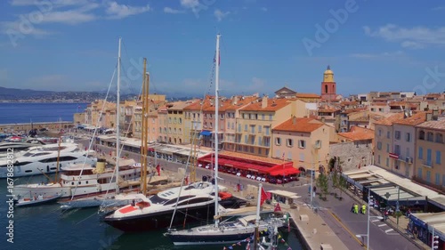panoramic aerial view captures the vibrant Port of Saint Tropez, showcasing luxurious yachts and sailboats docked alongside the colorful old town houses under a clear blue sky.
