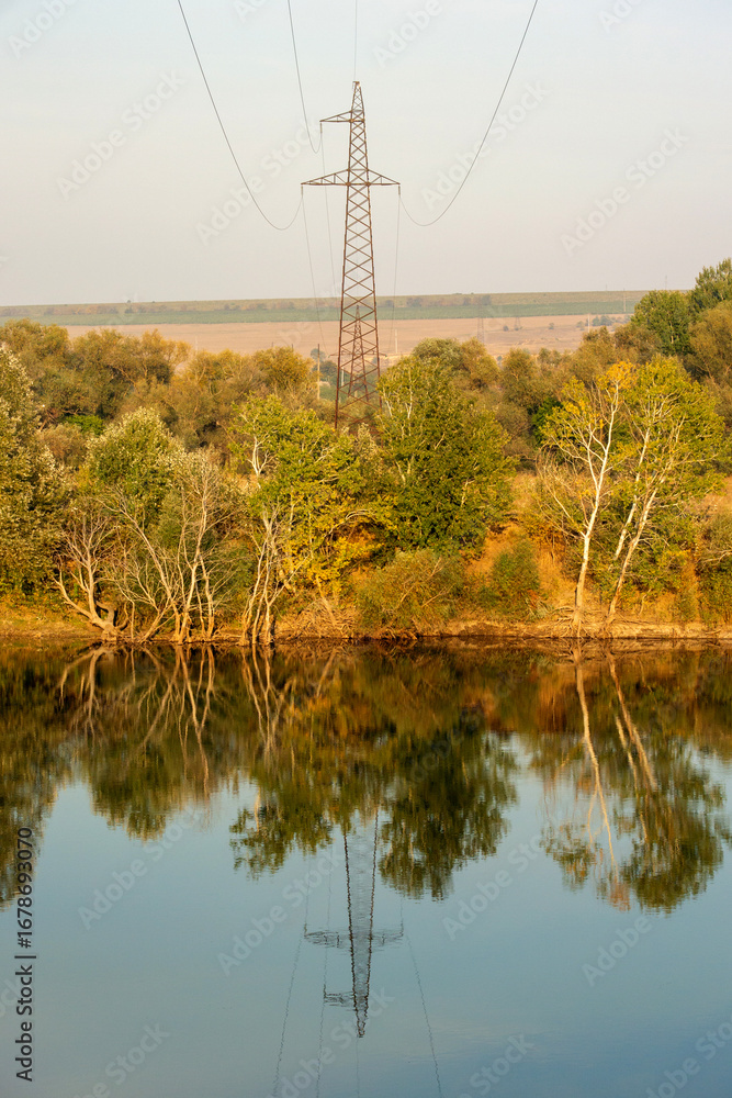 Obraz premium A serene, balanced landscape featuring a power line tower reflected in a calm body of water. The composition perfectly blends industrial elements with the tranquil natural scenery for a modern, reflec