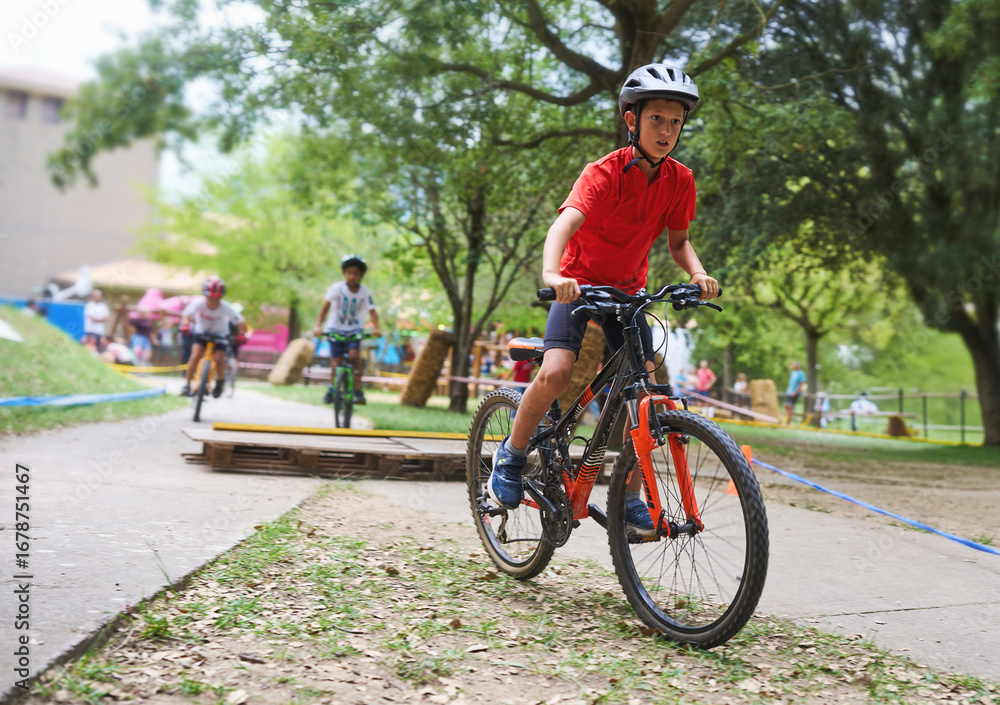 Fototapeta premium Focused boy in red shirt riding bicycle on park track with friends, enjoying outdoor sports, competition and childhood adventure