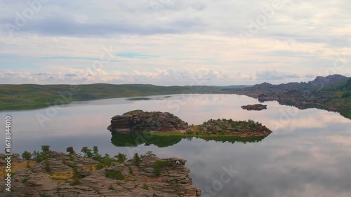 Tranquil lake with cloud reflections and rocky islands against green hills.