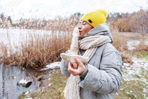 Middle aged woman in scarf and beanie warming hands by blowing on mittens in cold winter weather