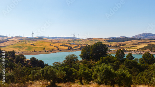 Panoramic view of a rural landscape in Andalusia, with golden hills and a turquoise reservoir under a clear blue sky. The summer sun illuminates the landscape and a wind farm in the distance, adding a