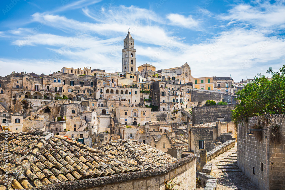 Obraz premium Daytime panoramic view of the historic town of Matera in southern Italy. The image highlights ancient stone houses, narrow streets, and the iconic Matera Cathedral rising above the Sassi cave dwelling
