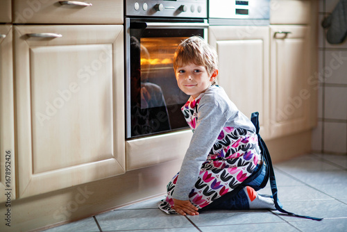 Cute blond boy baking muffins or pie in domestic kitchen. Happy child having fun with helping, sitting near ofen and waiting for cupcakes. Kid baking for Thanksgiving or Christmas holiday.