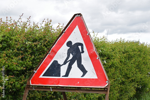 Close Up of Triangular Metal Traffic Sign with Workman Logo seen against Green Hedge