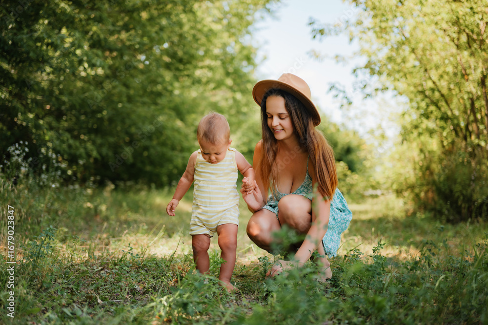 Fototapeta premium A smiling young mother in a summer dress and hat holds her little boy's hand as he takes his first steps in a grassy park on a sunny day.