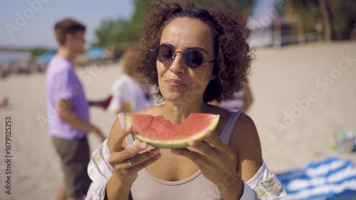 Young woman eating a slice of watermelon on the beach, with a group of friends in the background.