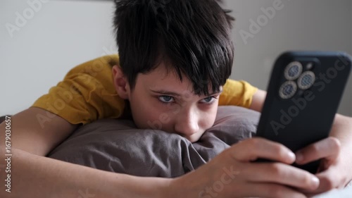 Closeup of a tired teenage boy lying on a pillow and watching a smartphone screen in a quiet bedroom at home.