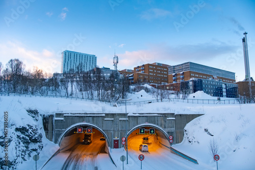Vehicles navigate a snowy tunnel in Tromso, Norway, surrounded by a winter landscape and urban architecture. Snow-covered trees and buildings frame the scene in serene twilight