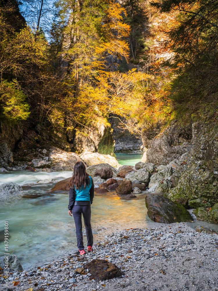 A woman stands on a rocky shore, gazing at a turquoise river flowing through a gorge with autumn foliage. Slizzya gorge, Julian Alps, Tarvisio, Ital A woman stands on a rocky shore, gazing at a turquoise river flowing through a gorge with autumn foliage. Slizzya gorge, Julian Alps, Tarvisio, Ital