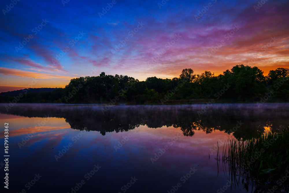 Obraz premium Sunrise lakeshore autumn landscape with forest, colorful clouds, and dawn sky reflected on pond water, a tranquil glimpse of Connecticut’s natural beauty in the heart of New England.
