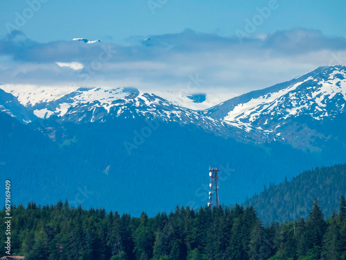 Cell tower rises above tree line of temperate rainforest near a building (bottom left) along Auke Bay, with view of snow-capped mountains and low clouds, on an afternoon in mid June, southeast Alaska