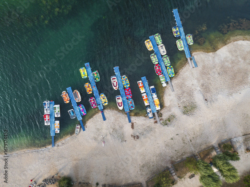 Aerial view of vibrant boats and pedalos tethered to blue jetties alongside a sandy beach meeting the clear turquoise waters of Lake Scanno, Scanno, Abruzzo, Italy.