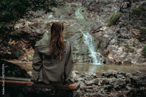 Young woman with long hair stands with her back to the camera, gazing at a mountain waterfall. The scene evokes freedom, serenity, and connection with nature.