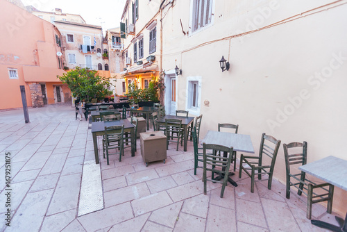 Empty taverna terrace with wooden chairs and tables in picturesque Mediterranean square surrounded by traditional pastel-colored architecture. Corfu, Greece.