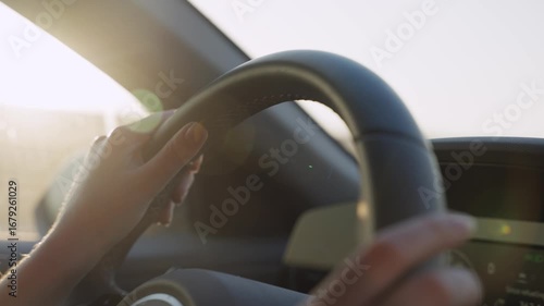 Female hands on car steering wheel driving on highway road with sun shining. Close up on wheel as woman holding it commuting to work or going on vacation or escaping. Sunset or sunrise in background