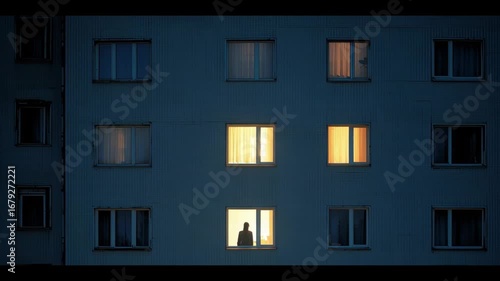 La imagen muestra un edificio iluminado por la noche, destacando ventanas con luces encendidas. Se pueden apreciar siluetas cerca de las ventanas. Una atmósfera tranquila y urbana.
