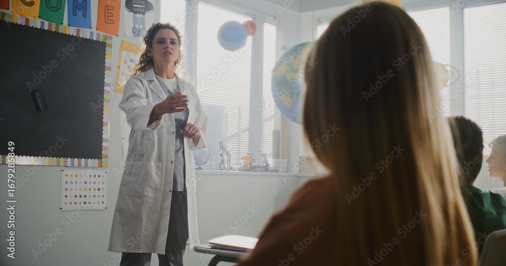 Fototapeta premium Female Doctor Coat Visiting Classroom, Giving Lecture to Curious Primary School Students About Health and Hygiene. Young Kids Sitting and Listening to Healthcare Professional. Basic Medical Education.