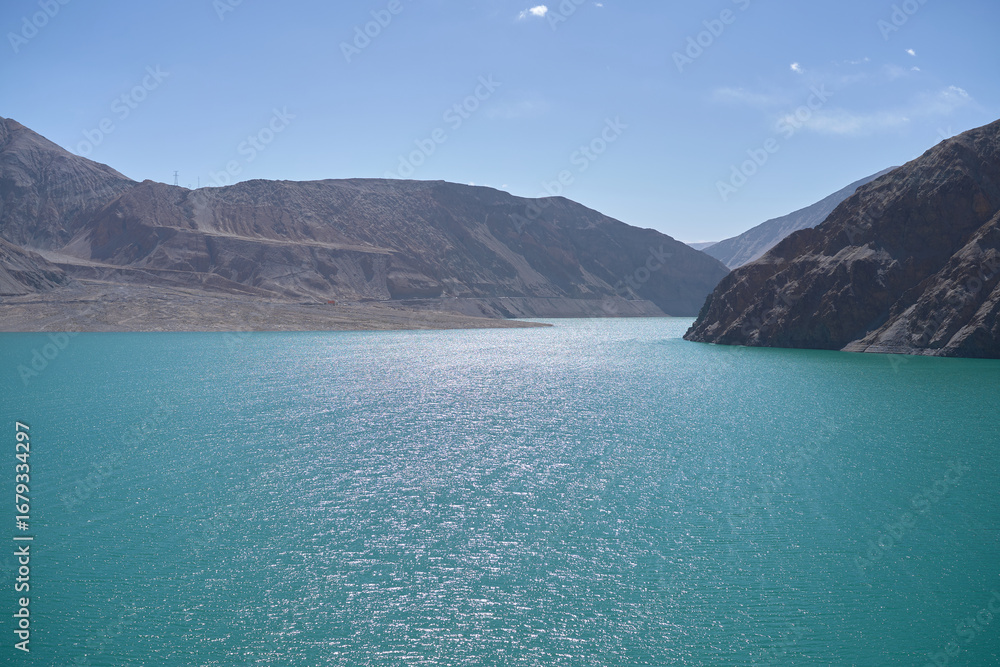 Obraz premium Sparkling Turquoise Water Reflecting Sunlight Amidst Rugged Mountain Landscape Under a Clear Blue Sky