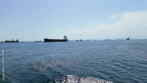 Numerous cargo ships float on tranquil waters nearby the coast, set against a backdrop of clear blue skies. The bright sun enhances the peaceful atmosphere of the scene.