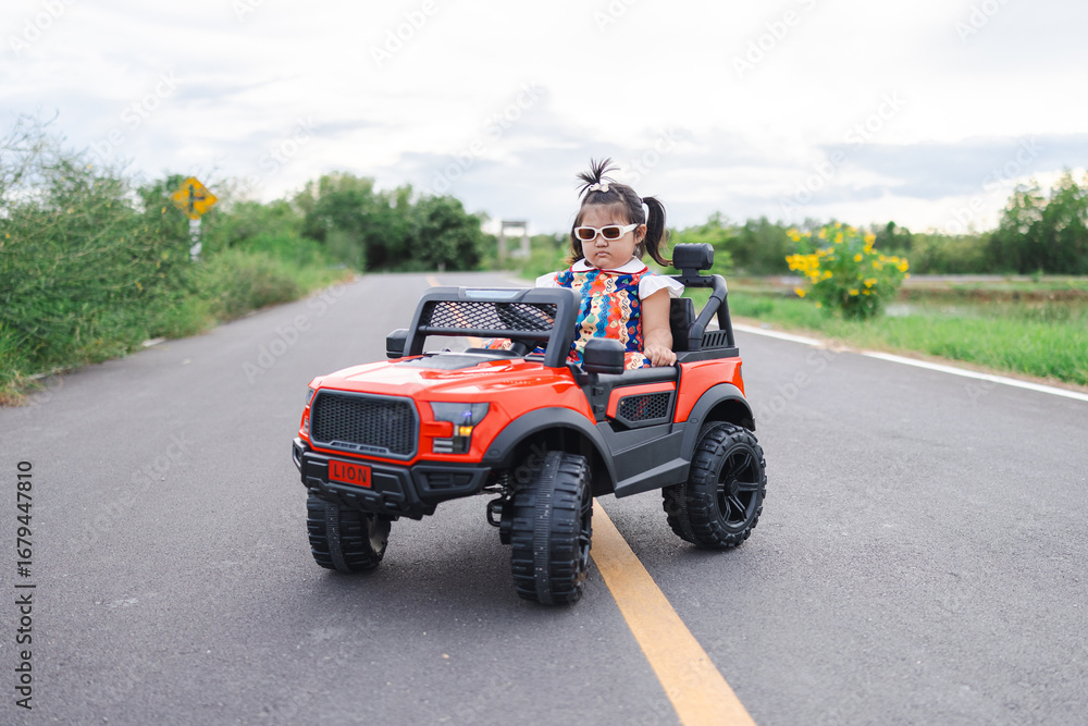 Fototapeta premium A cute little Asian girl is happily riding a small toy car along a country road,A girl drives a toy car on a concrete road.