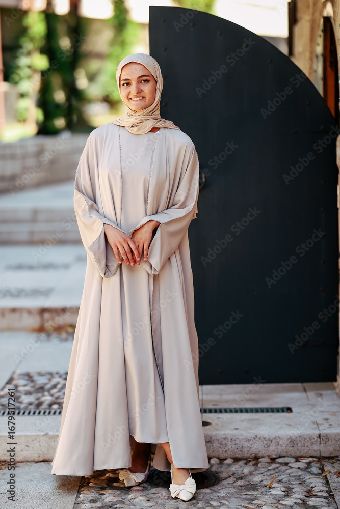 Naklejka premium Smiling woman in modest hijab and flowing dress adjusting scarf outdoors, standing near stone steps, symbolizing elegance, culture and modern lifestyle.