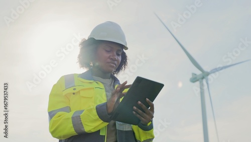 Focused African American woman holding tablet device while tapping on screen with one finger. Recording her observations from checking wind turbine power plant. Protective equipment.