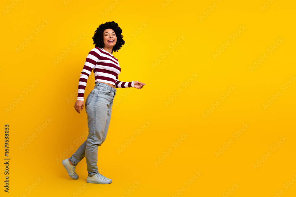 Fototapeta premium Confident young woman in striped top walking confidently against yellow background showcasing casual modern style