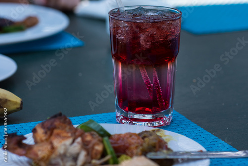 grilled chicken breast, A glass with a dark red drink and a straw. It contains ice cubes and possibly berries that give the drink its rich color. Next to the drink, on a plate, there is food