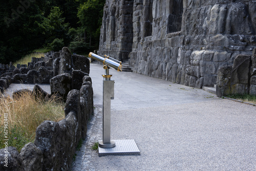 A modern metallic telescope stands near the historic stone wall, offering visitors a panoramic view of the surrounding landscape