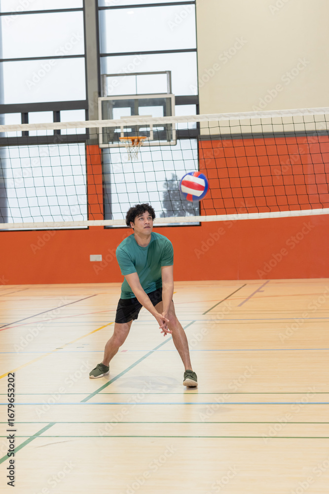 Obraz premium Teenage male volleyball athlete in shirt passing volleyball in gym under net with basketball hoop