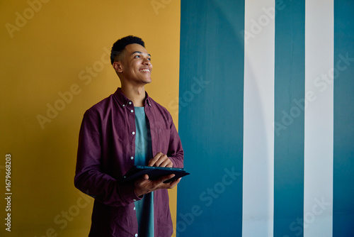 Smiling young businessman looking away while using tablet against colorful wall in office