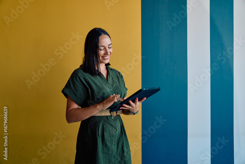 Smiling young businesswoman in smart casual using tablet against colorful wall in office
