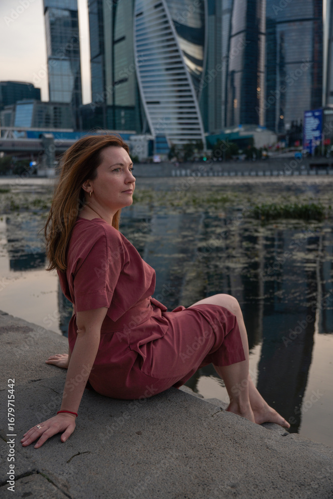 Fototapeta premium Woman in a red dress sits by the Moscow River with the iconic skyscrapers of Moscow City in the background