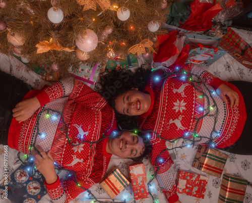 Latin couple lying on the floor with the Christmas tree next to them, smiling and wearing red Christmas sweaters, wide angle shot zenithal