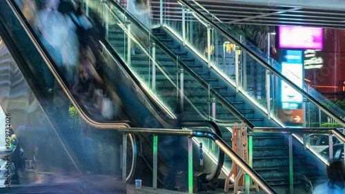 Blurred people ascend and descend a modern escalator in a busy urban environment at night. The long exposure photography technique captures the dynamic movement of the crowd, resulting in abstract