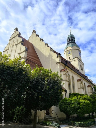 Beautiful view of a historic European church featuring a clock tower, stepped gables, and arched windows. Surrounded by lush greenery under a partly cloudy blue sky. Religious and cultural heritage.