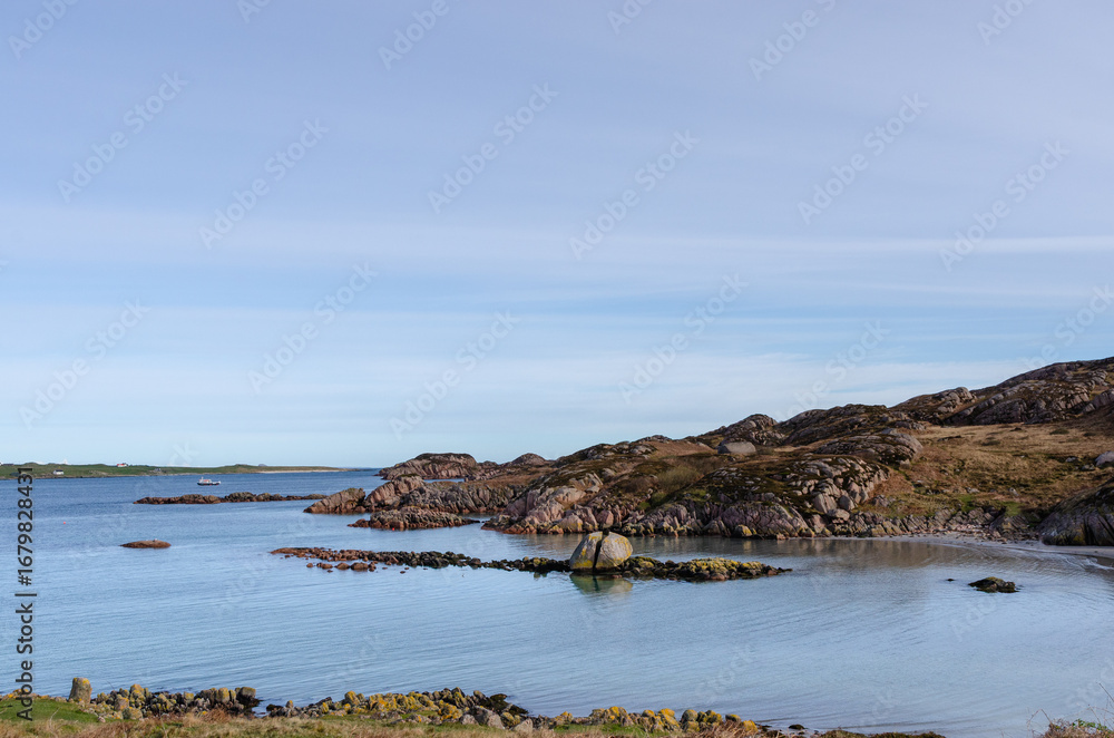 Fototapeta premium Rocky shoreline and blue coastal waters of the Isle of Mull, with distant green hills under a clear sky. A serene and natural island scene.